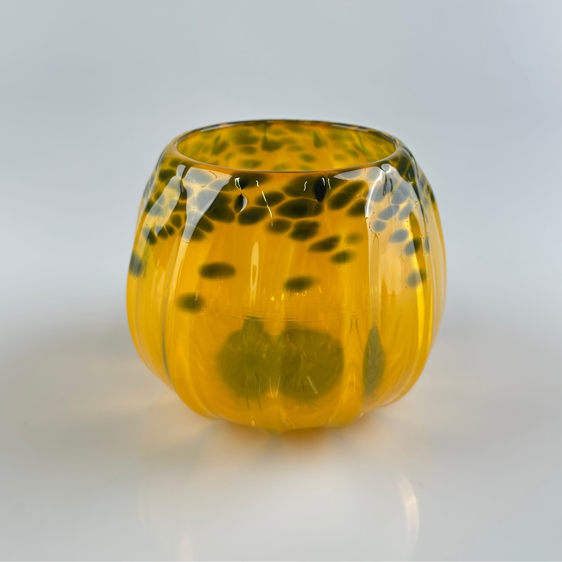 A small, rounded glass vessel with a ribbed pumpkin-like texture, featuring a translucent amber-yellow body with dark black mottled spots.
