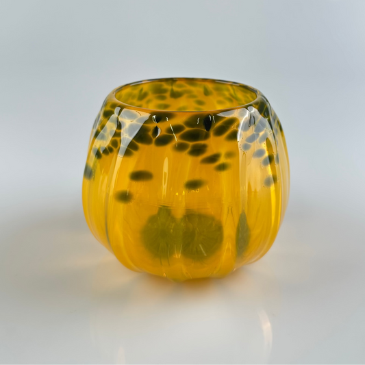 A small, rounded glass vessel with a ribbed pumpkin-like texture, featuring a translucent amber-yellow body with dark black mottled spots.