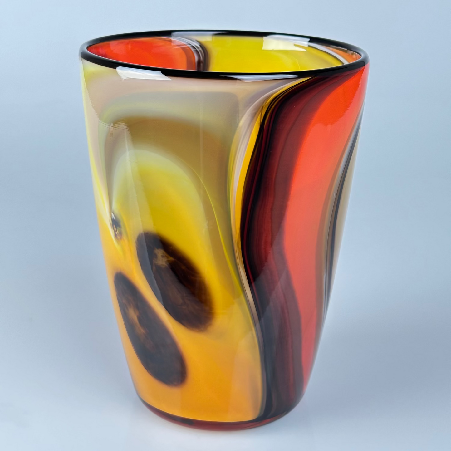 A hand-blown glass vase with a marbled swirl pattern of yellow, orange, red, and dark brown, set against a light gray background.