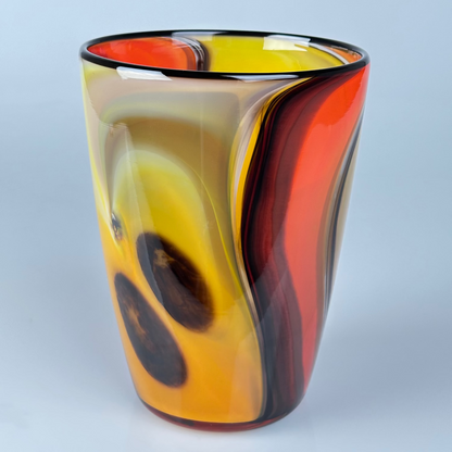 A hand-blown glass vase with a marbled swirl pattern of yellow, orange, red, and dark brown, set against a light gray background.